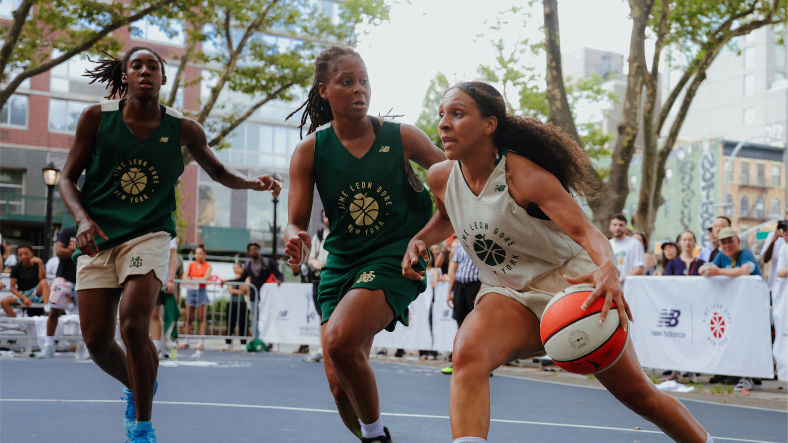 Three women playing basketball on an outdoor court with spectators in the background.