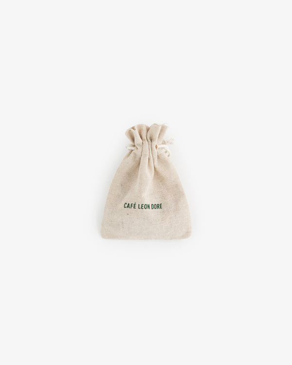 A small beige drawstring pouch for Café Leon Dore Komboloi, with branding on the front, centered on a white background.