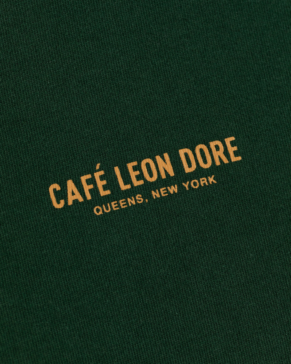Green Café Leon Dore Logo Tee with yellow “CAFÉ LEON DORE QUEENS, NEW YORK” text in uppercase letters.
