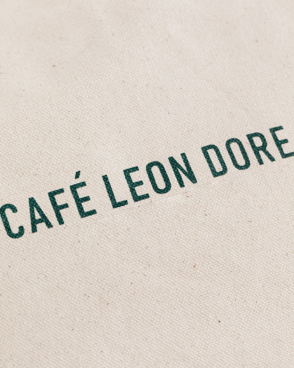The Café Leon Dore Tote Bag features green text on a textured, off-white fabric background.