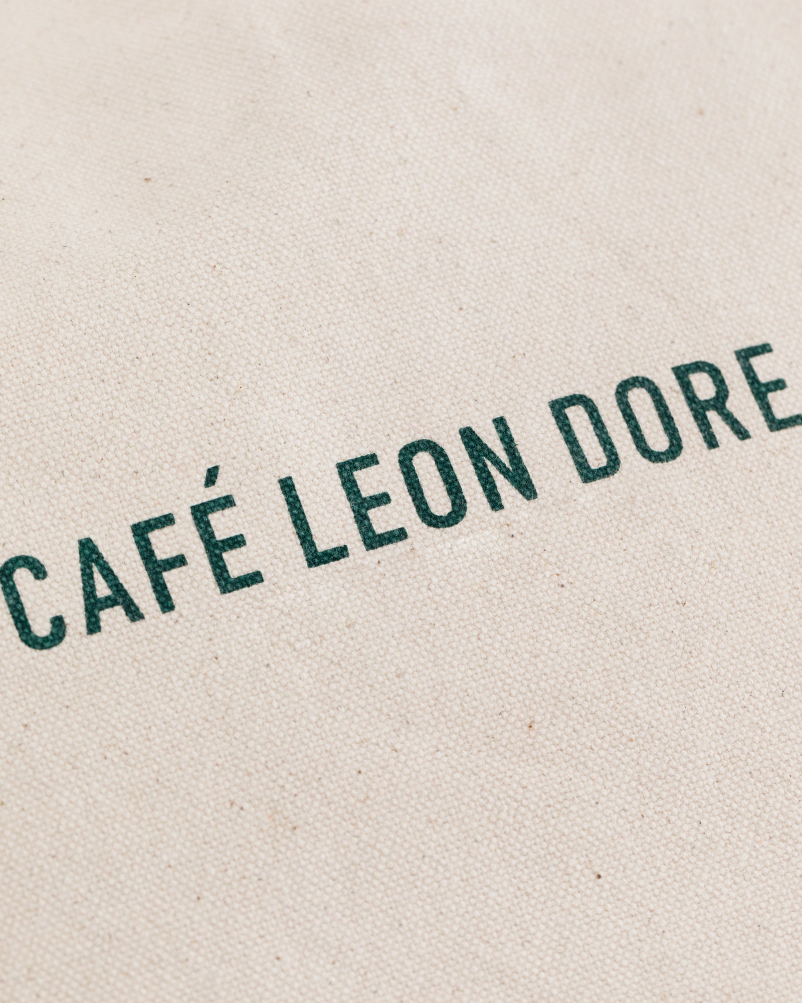The Café Leon Dore Tote Bag features green text on a textured, off-white fabric background.