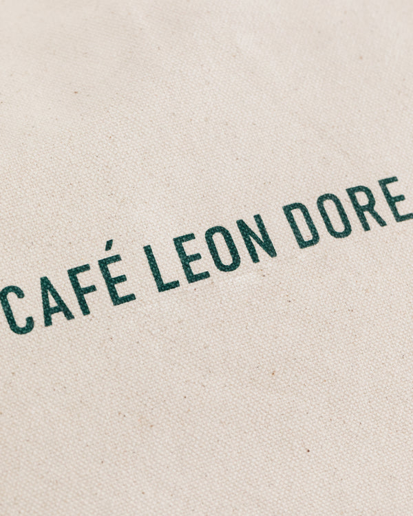The Café Leon Dore Tote Bag features green text on a textured, off-white fabric background.