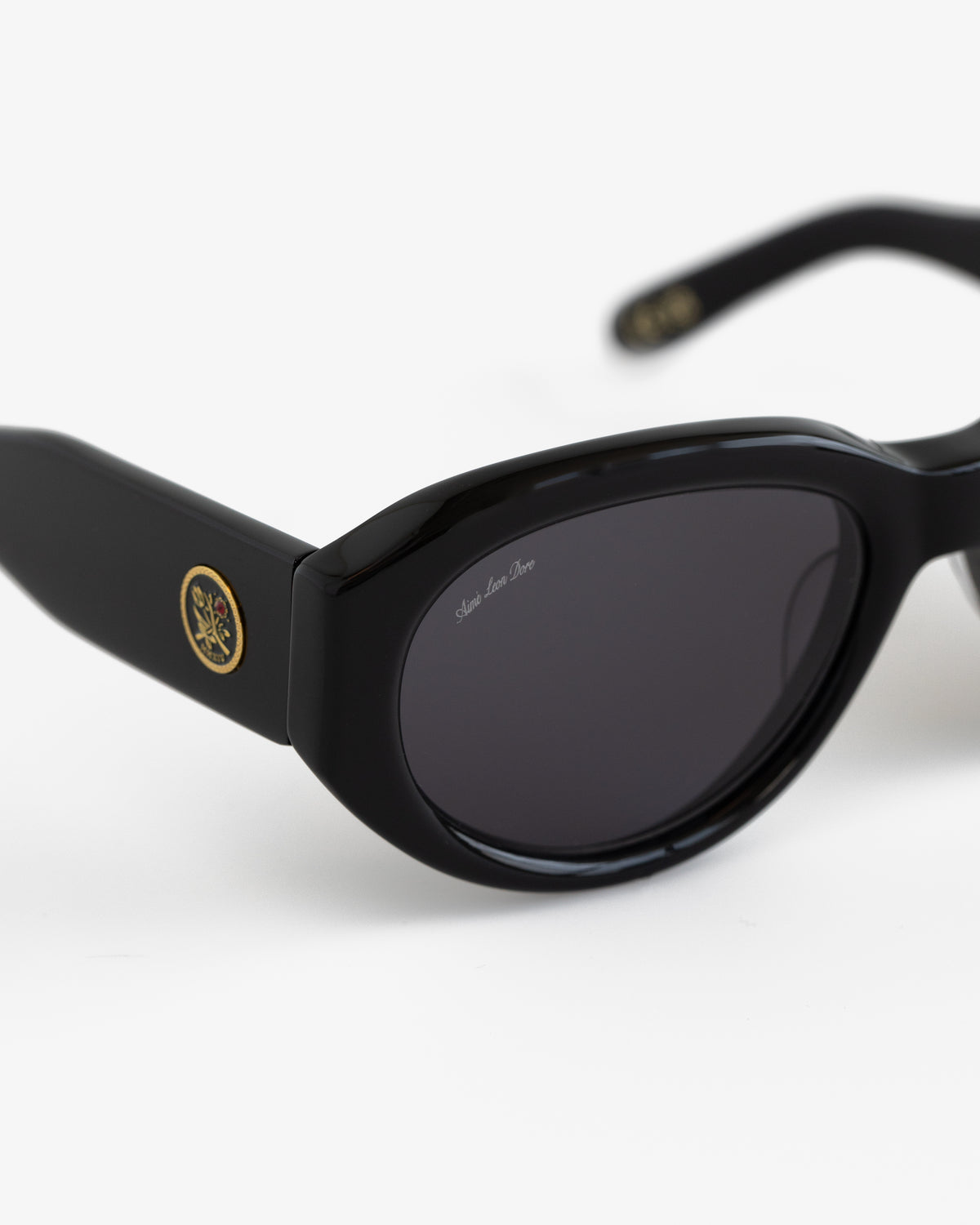 Jackie O Sunglasses: black oval frames, dark lenses, gold emblem on temple, shown on a white background.