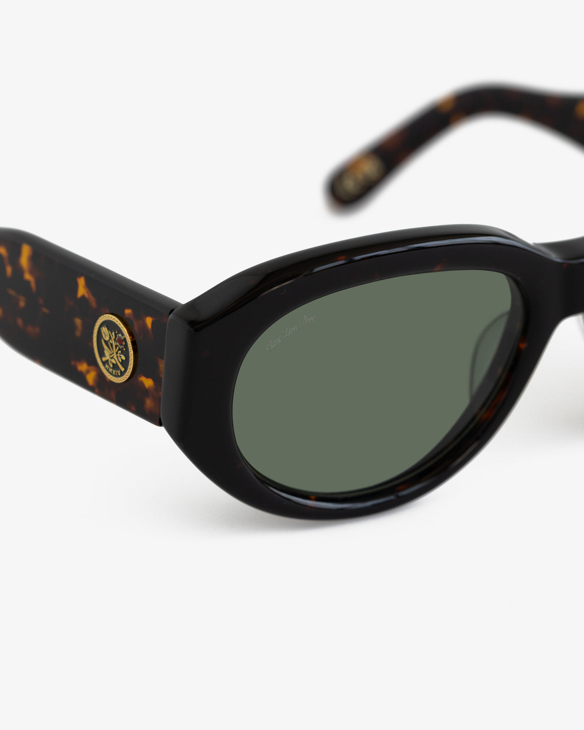 Close-up of Jackie O Sunglasses featuring tortoiseshell frames, dark green lenses, and a gold emblem on the temple.