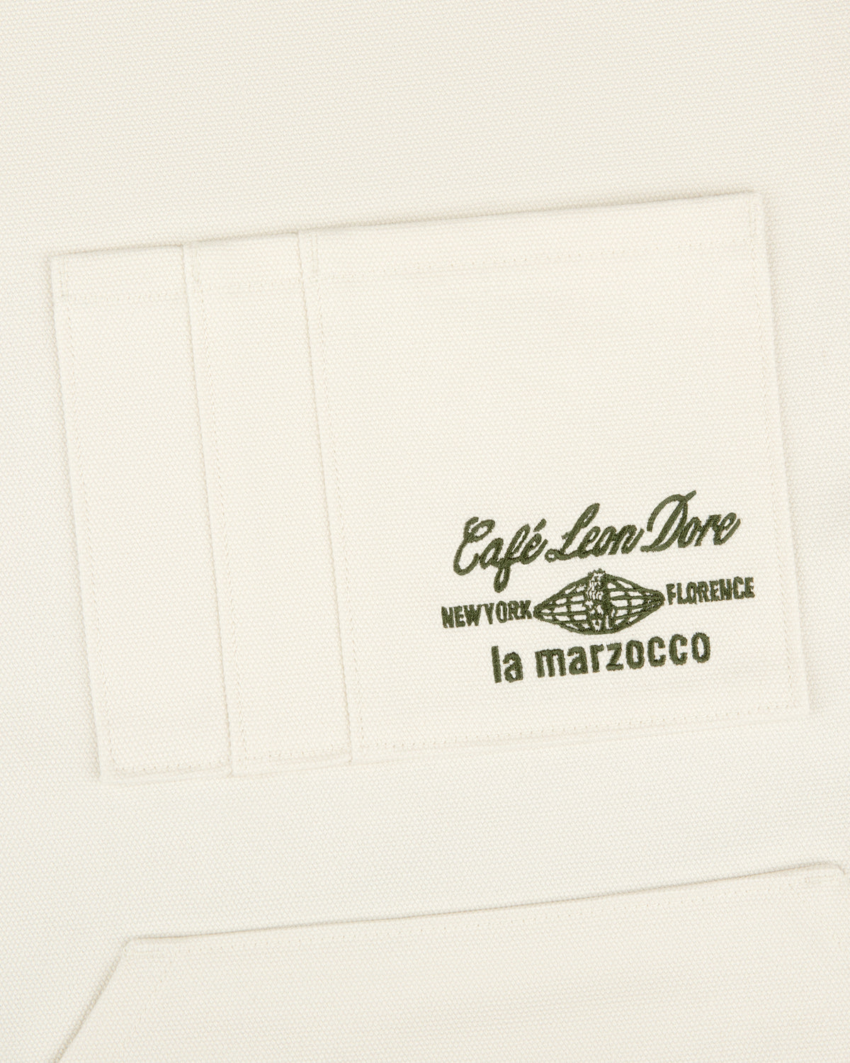White apron pocket with green text "Café Leon Dore / La Marzocco" and a coffee bean illustration.