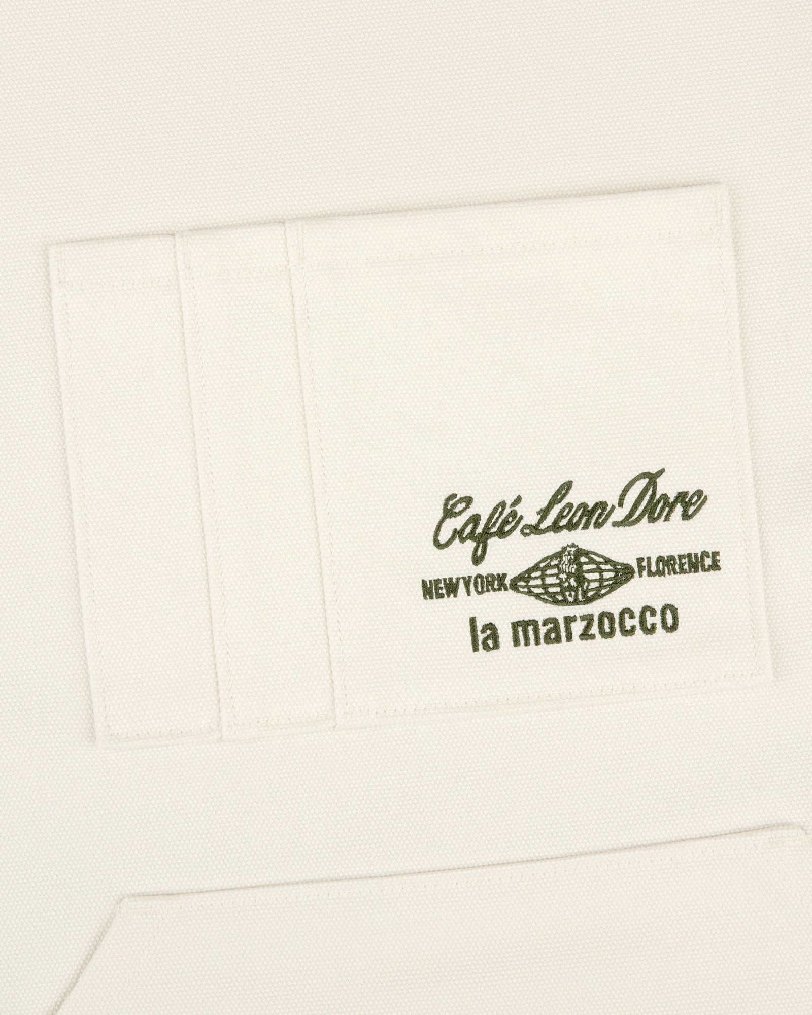 White apron pocket with green text "Café Leon Dore / La Marzocco" and a coffee bean illustration.