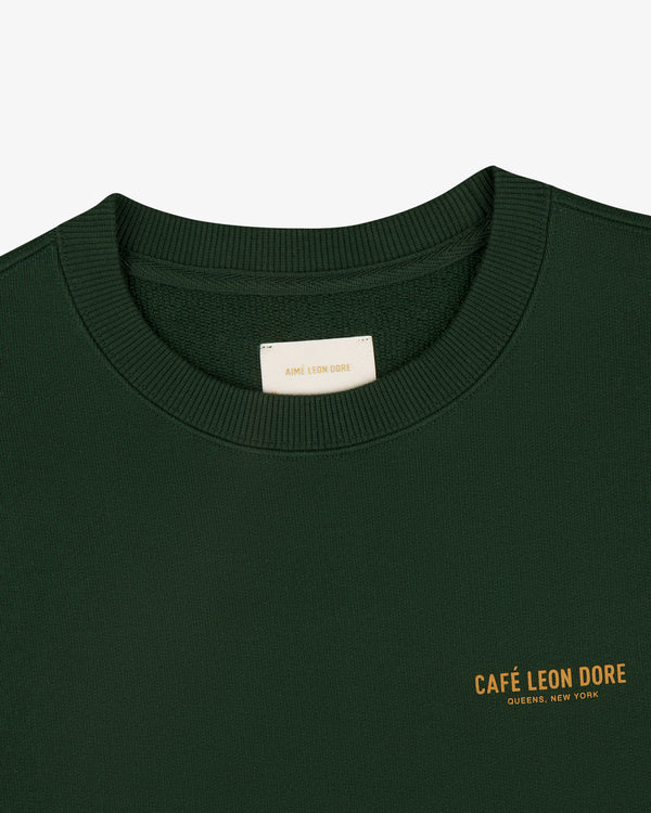 Café Leon Dore Crewneck Sweatshirt in dark green with yellow "CAFÉ LEON DORE QUEENS, NEW YORK" chest print.