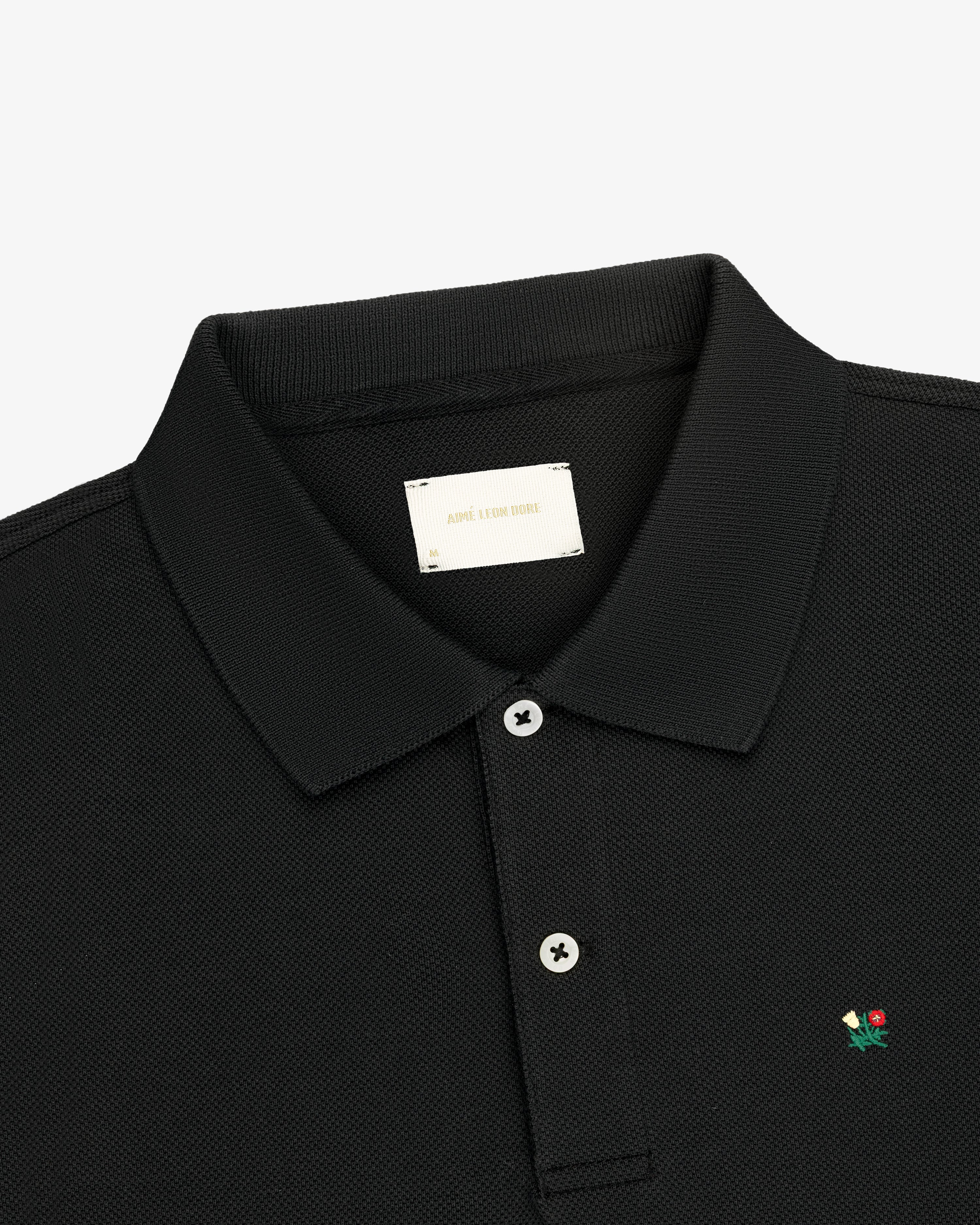 Crest Piqué Polo: black, collared, two buttons, small flower logo embroidered on the chest.