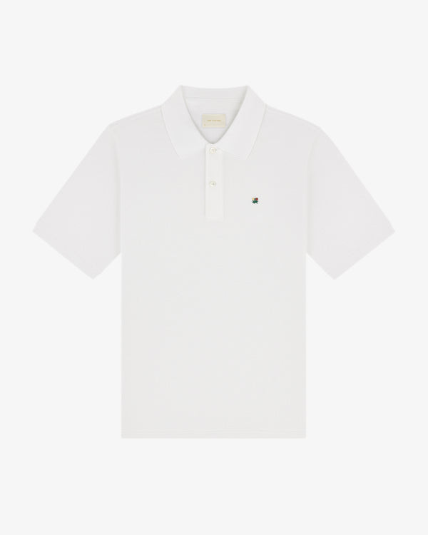 The Crest Piqué Polo is a white short-sleeve shirt with a collar and small embroidered logo on the left chest.