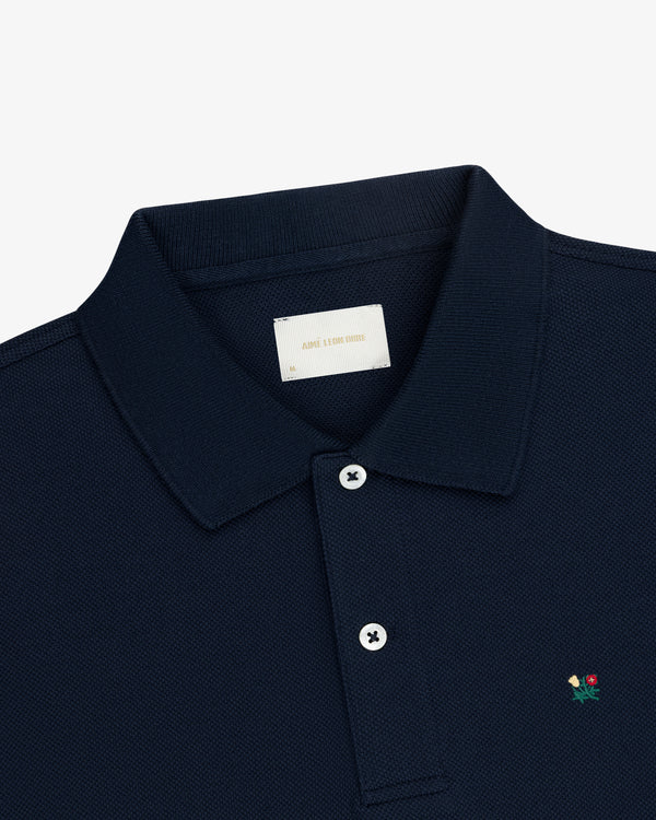Crest Piqué Polo in navy blue with a small embroidered flower and two-button placket, shown on a white background.