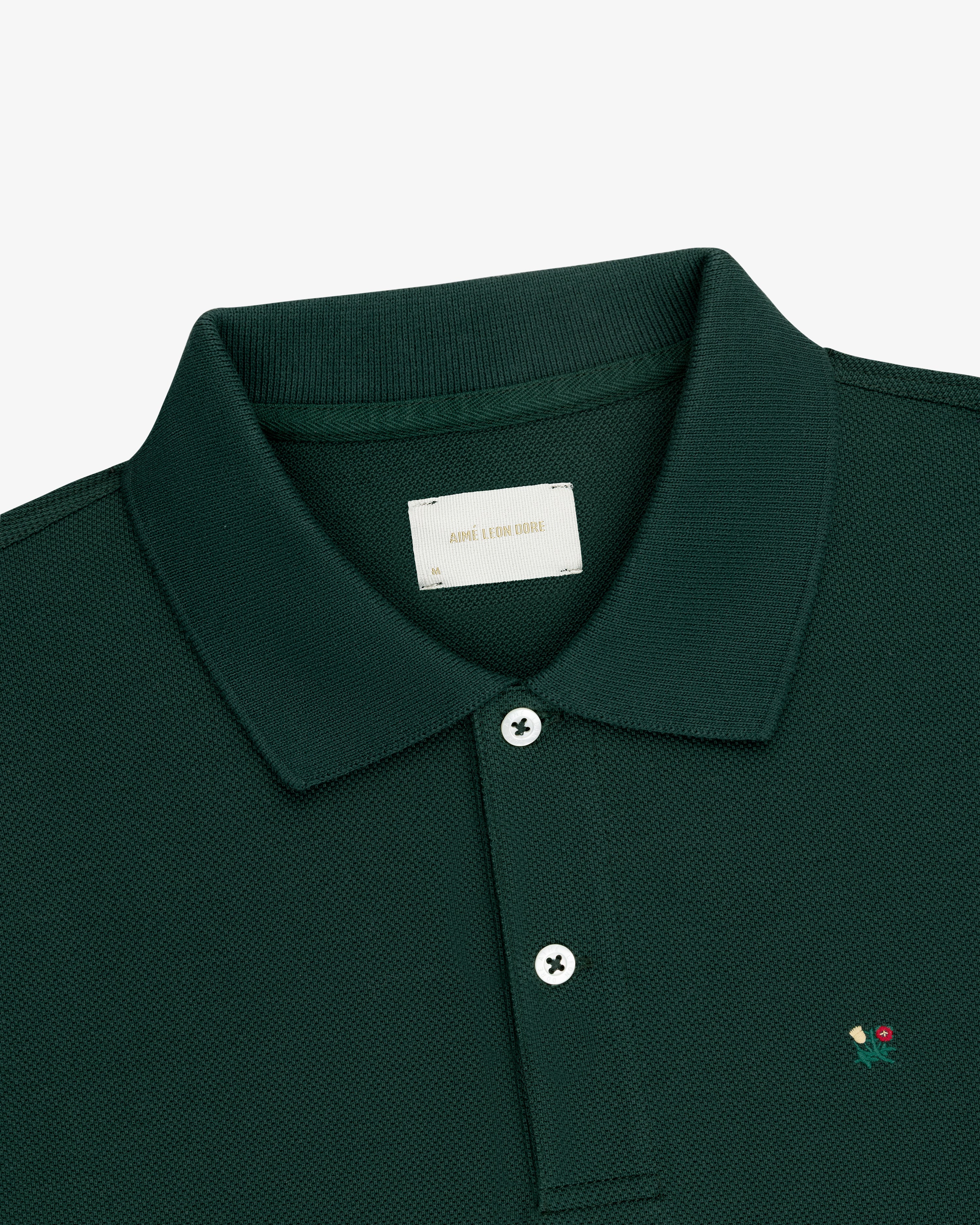 Crest Piqué Polo in dark green with a collar, two white buttons, and red & yellow chest embroidery.