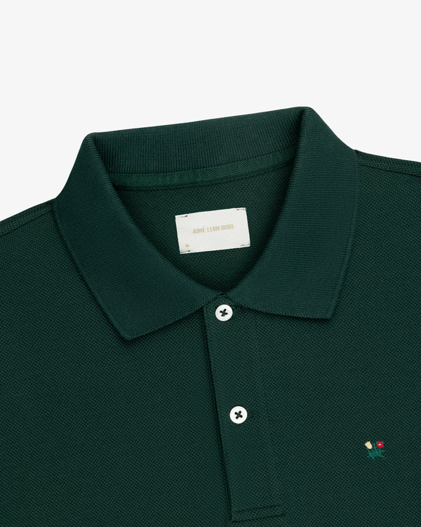 Crest Piqué Polo in dark green with a collar, two white buttons, and red & yellow chest embroidery.