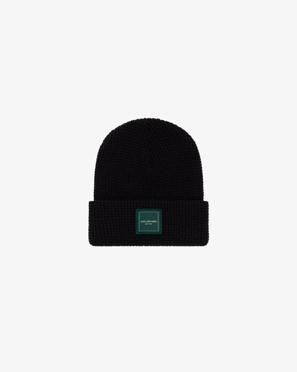 Café Leon Dore Beanie: black knit with folded brim and rectangular green patch, shown on a white background.