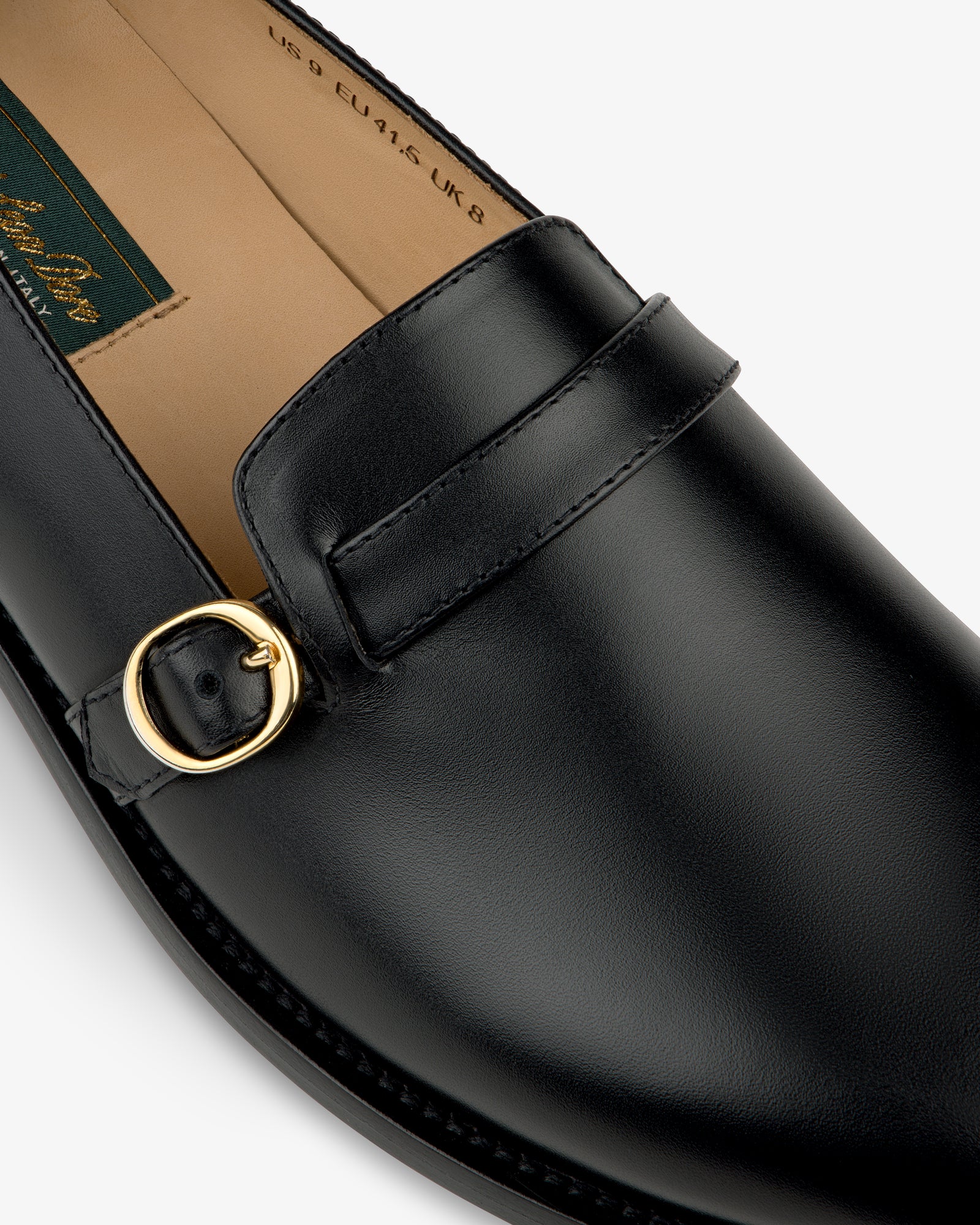 Close-up of the Venetian Buckle Loafer in black leather with gold buckle and visible stitching on a white background.