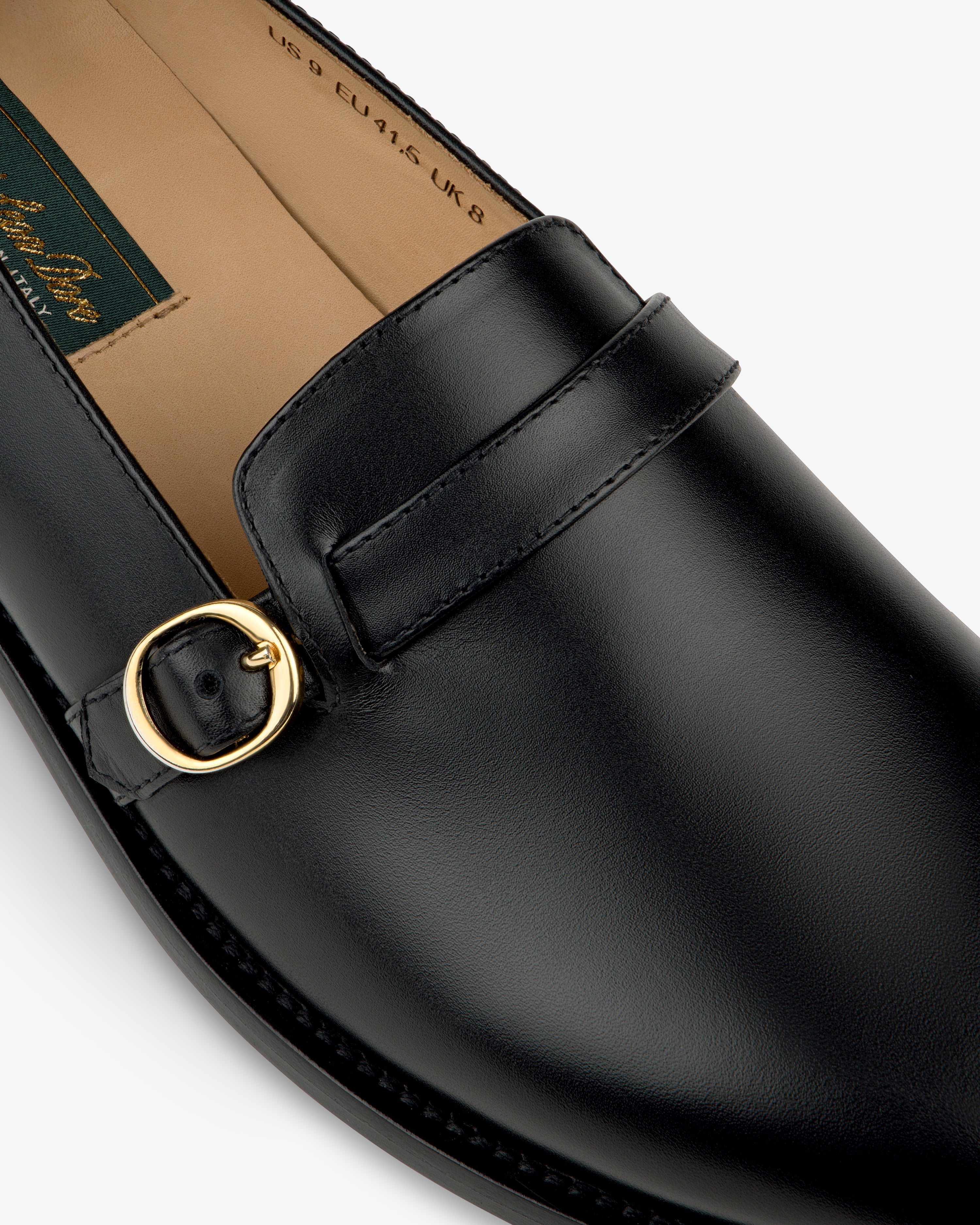 Close-up of the Venetian Buckle Loafer in black leather with gold buckle and visible stitching on a white background.