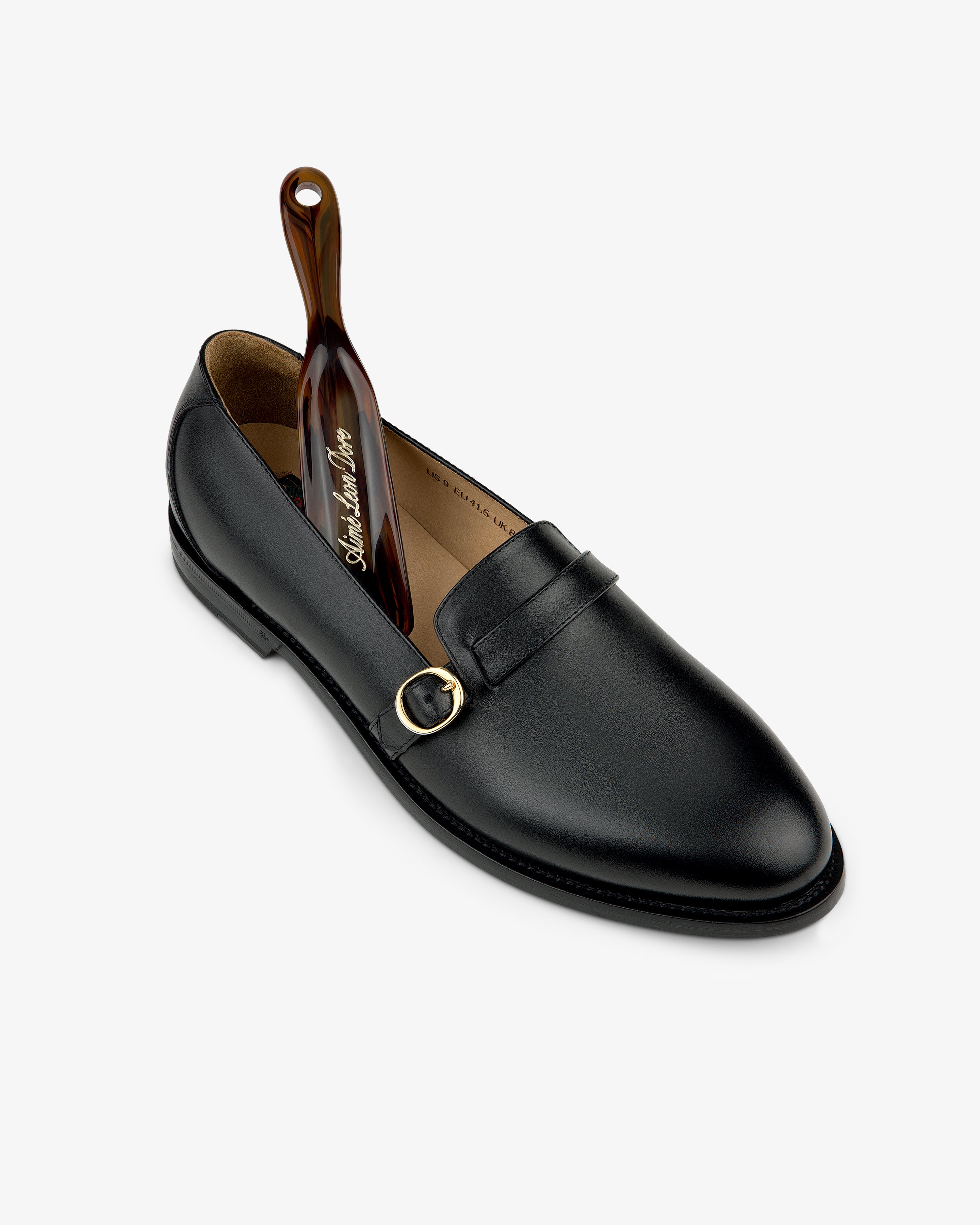 Venetian Buckle Loafer in black leather with gold buckle and brown shoehorn, displayed on a white background.