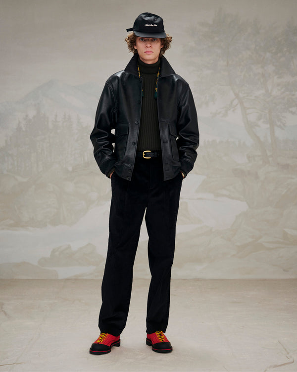 Person styled in a black leather jacket, black pants, and Suede Derby Hiker shoes before a muted landscape backdrop.