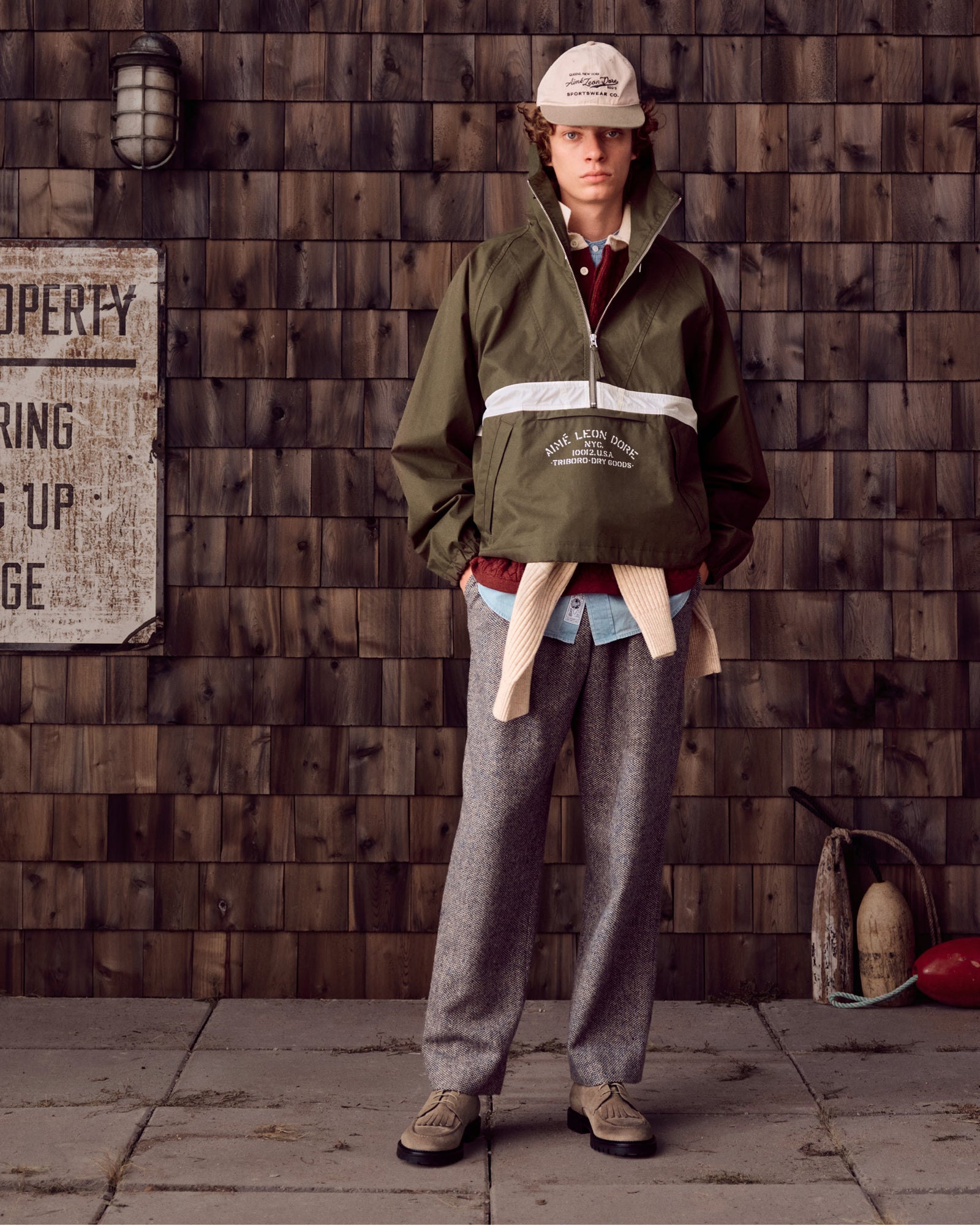 Person wearing Suede Lug Sole Derby, green jacket, gray trousers, and cap stands by a shingle wall with vintage props.