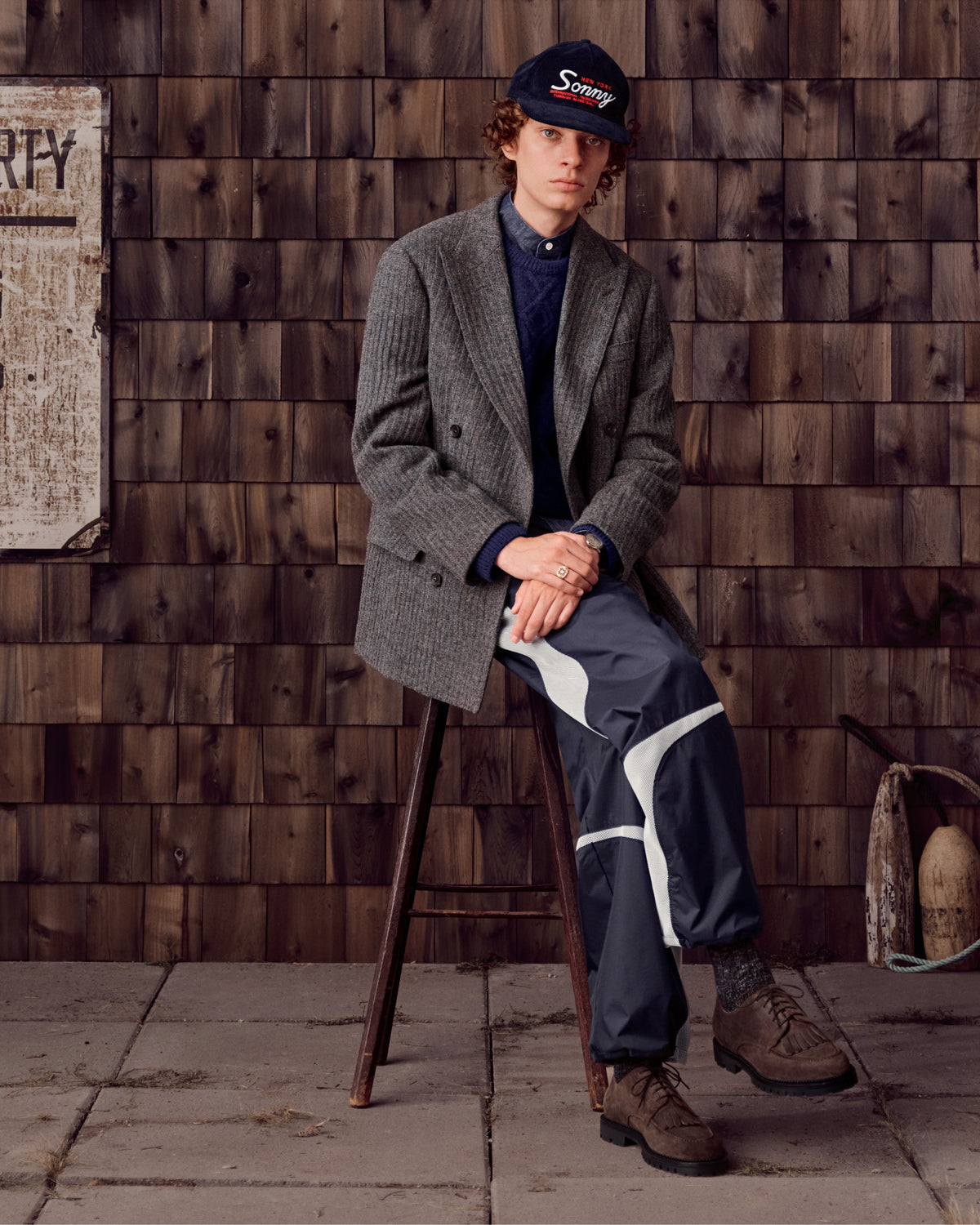 A person in Suede Lug Sole Derby shoes sits on a stool against a wooden wall background.