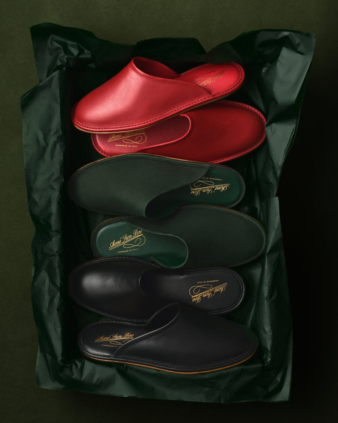 Three pairs of Nubuck Travel Slippers—red, green, and black—on green tissue paper with a dark green background.