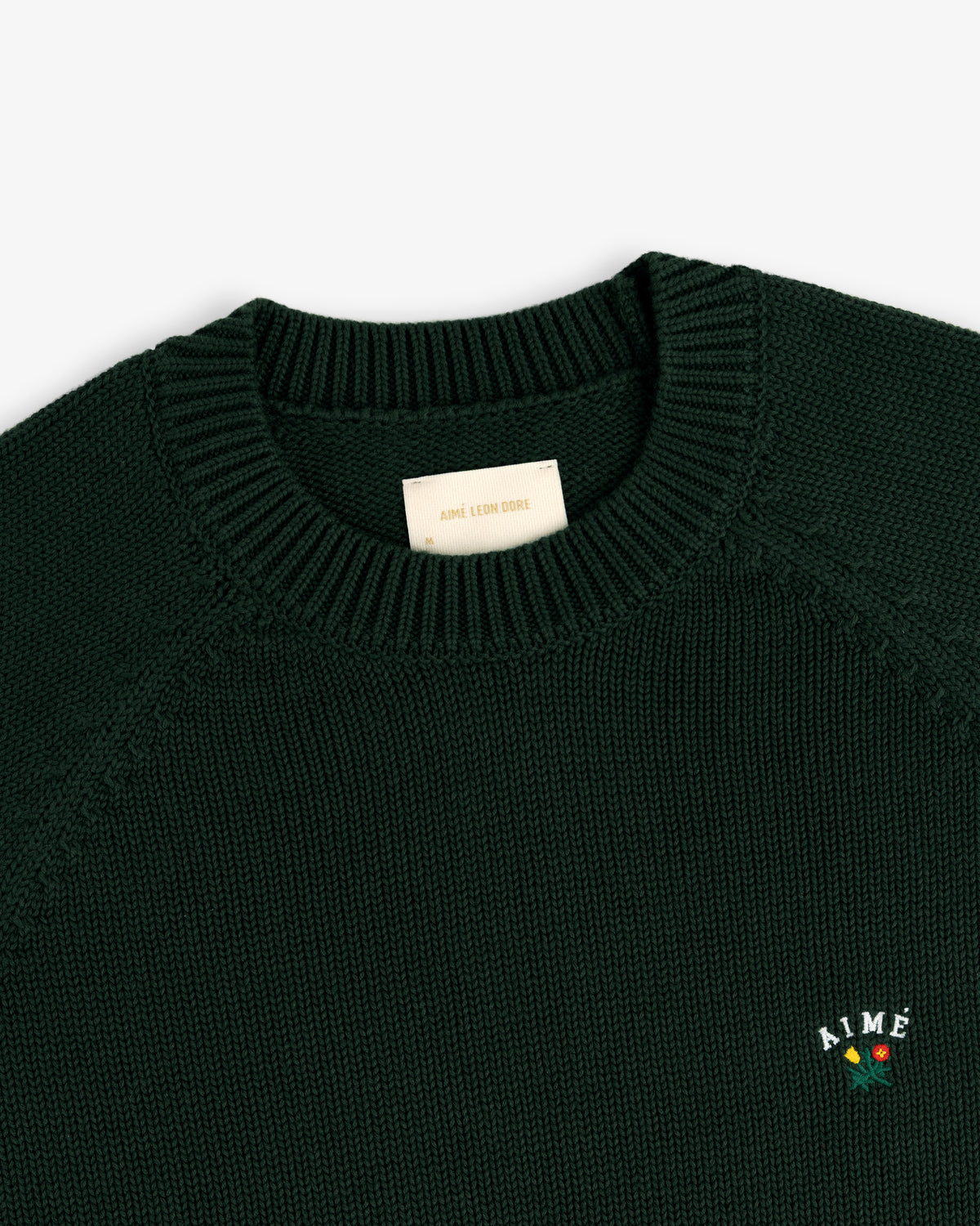 Crest Raglan Sweater in dark green with AIMÉ and rose embroidery on the left chest, shown on white background.