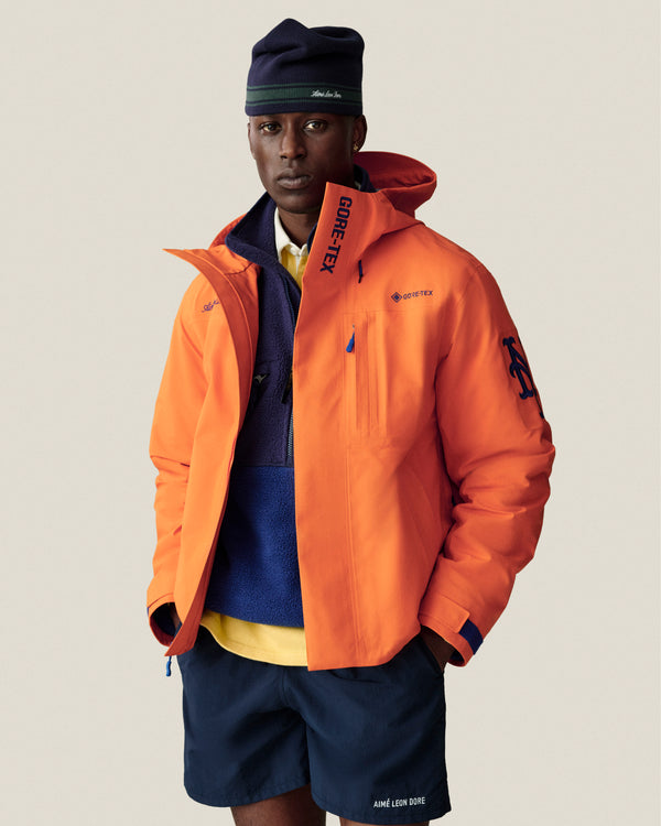 A man wears a navy beanie, Mets GORE-TEX Rain Delay Jacket, blue fleece, yellow shirt, and navy Aimé Leon Dore shorts.