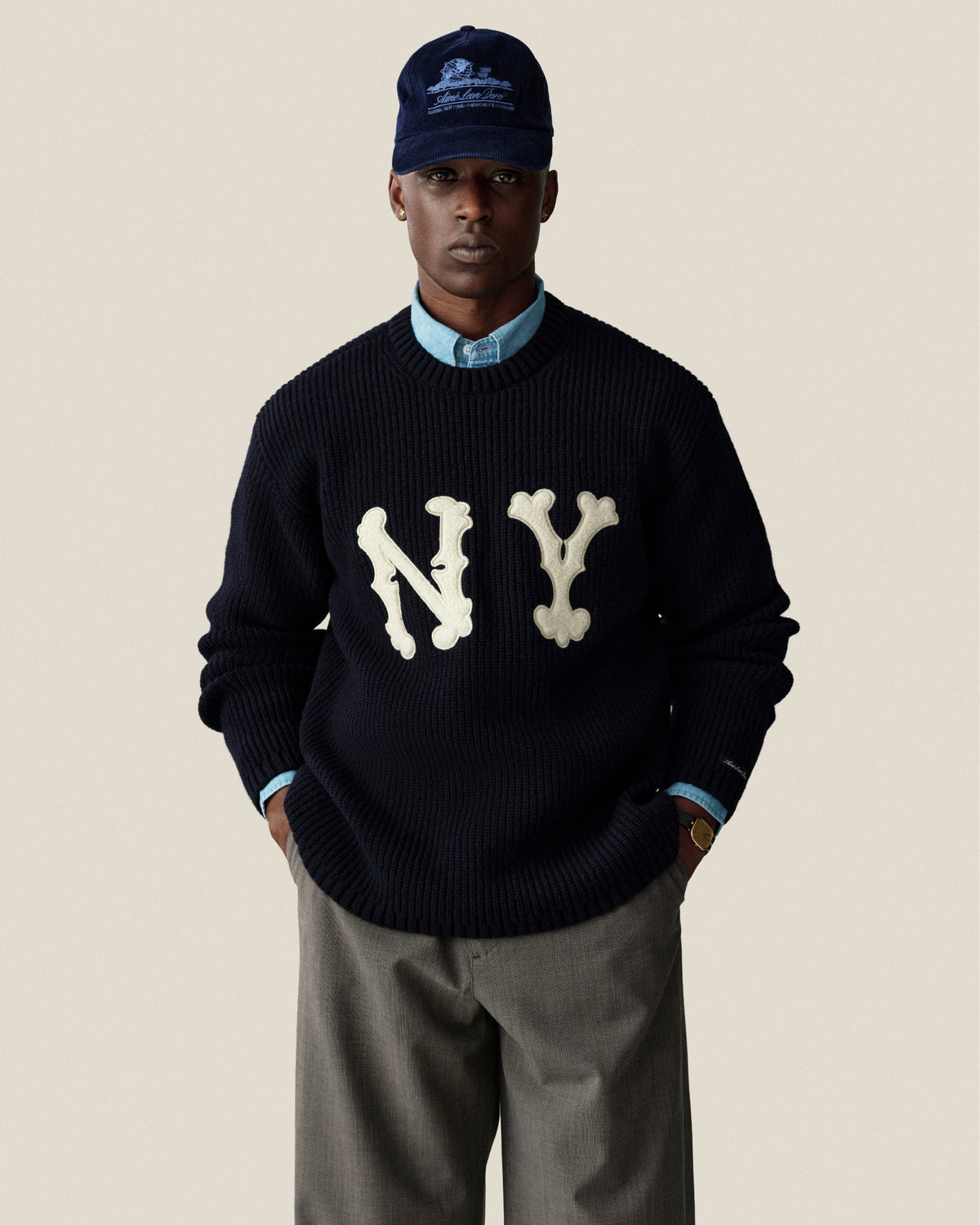 A person wears a navy NY sweater, blue shirt, Houndstooth Elasticated Waist Trouser, and navy cap against a plain background.