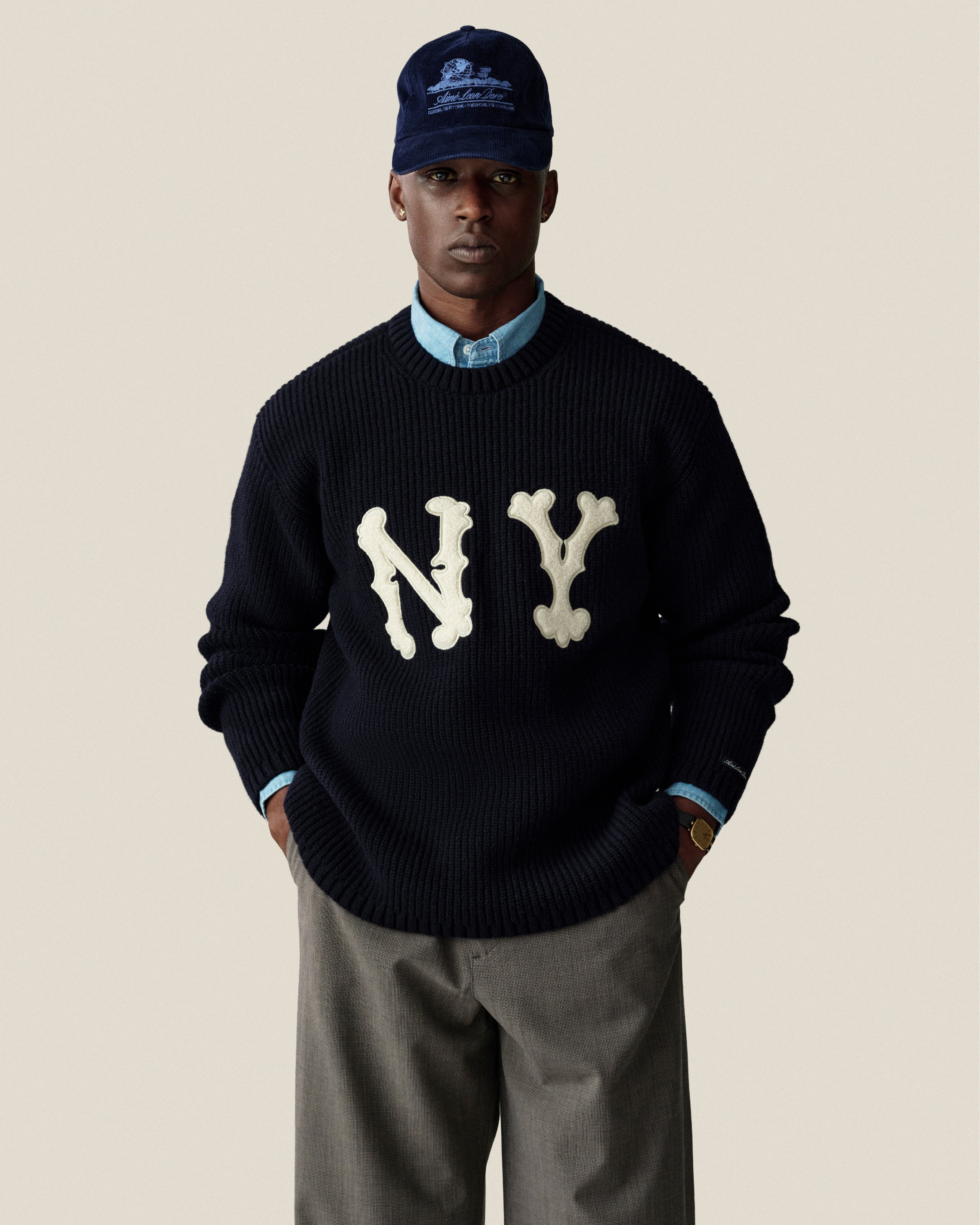 A person wears a navy NY sweater, blue shirt, Houndstooth Elasticated Waist Trouser, and navy cap against a plain background.