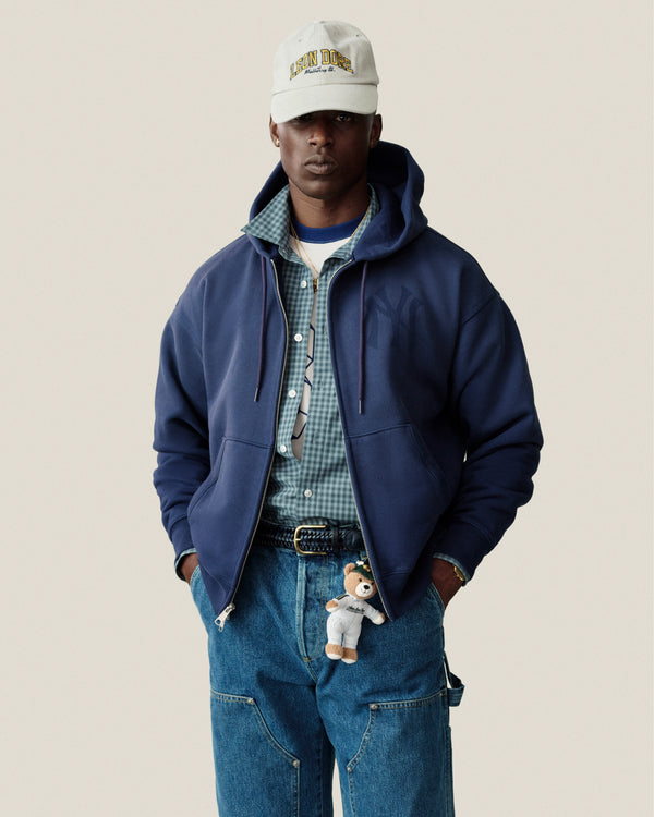 Man in Yankees Washed Full-Zip Hoodie, plaid shirt, blue jeans, cap, and teddy bear keychain against plain background.