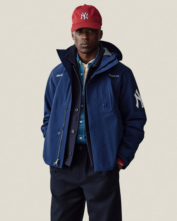 Man wearing a Yankees GORE-TEX Rain Delay Jacket and red Yankees cap, standing with hands in pockets.
