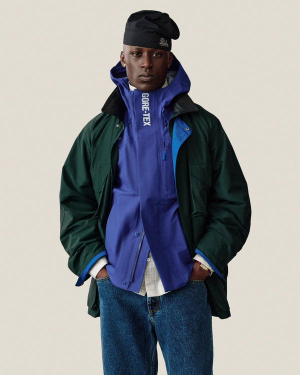 A person wears a Unisphere Skull Cap, green jacket, blue GORE-TEX jacket, and blue jeans while facing forward.