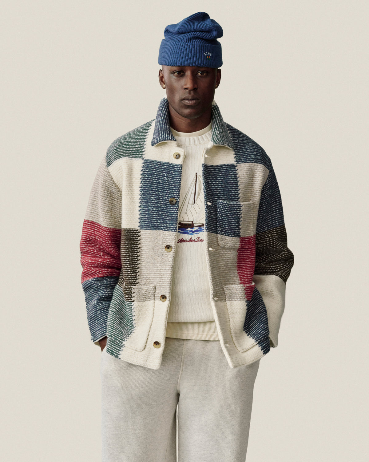 Man in blue beanie, patchwork jacket, sailboat sweater, and Stone Washed Marina Sweatpants stands against plain background.