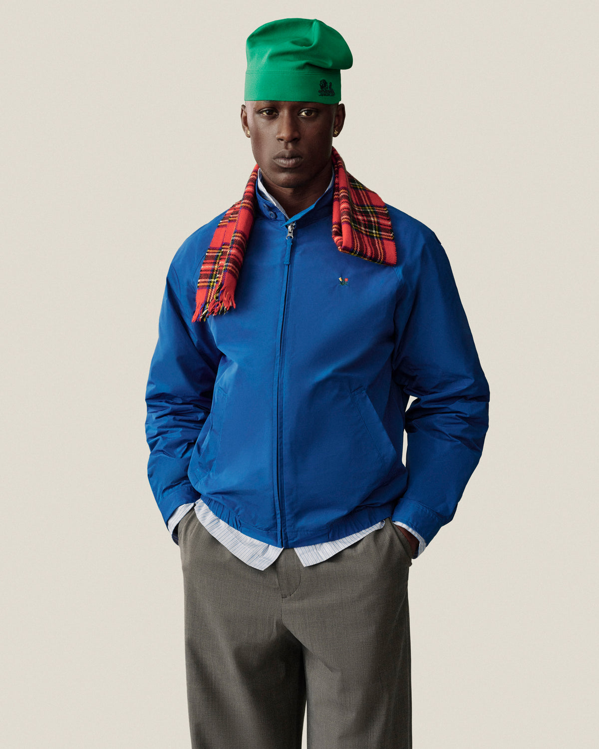 Man in blue jacket, green hat, and red plaid scarf poses in Houndstooth Elasticated Waist Trouser against plain backdrop.