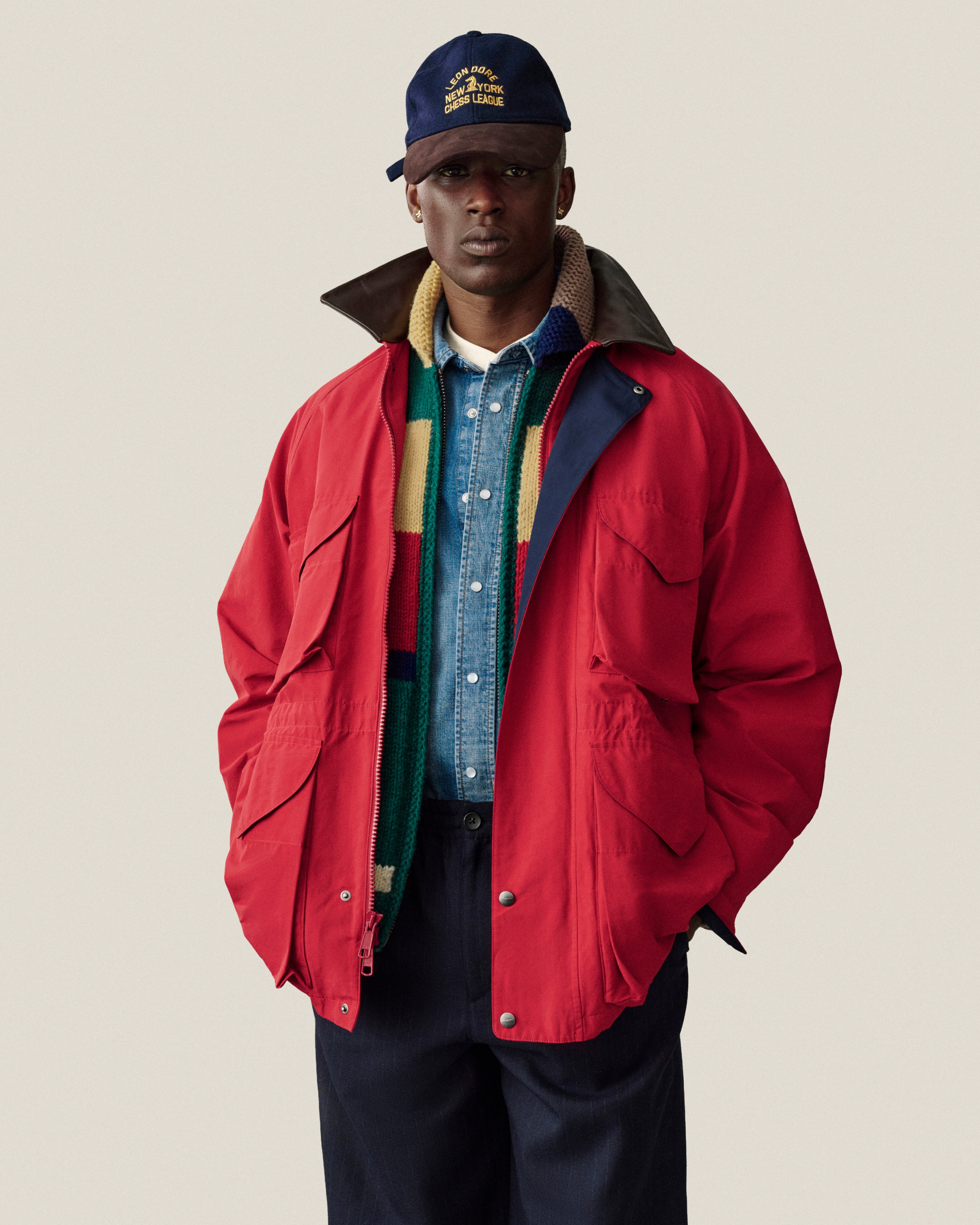 A man in a red jacket and scarf, wearing a Triborough Chess Club Wool Hat, stands with hands in pockets.