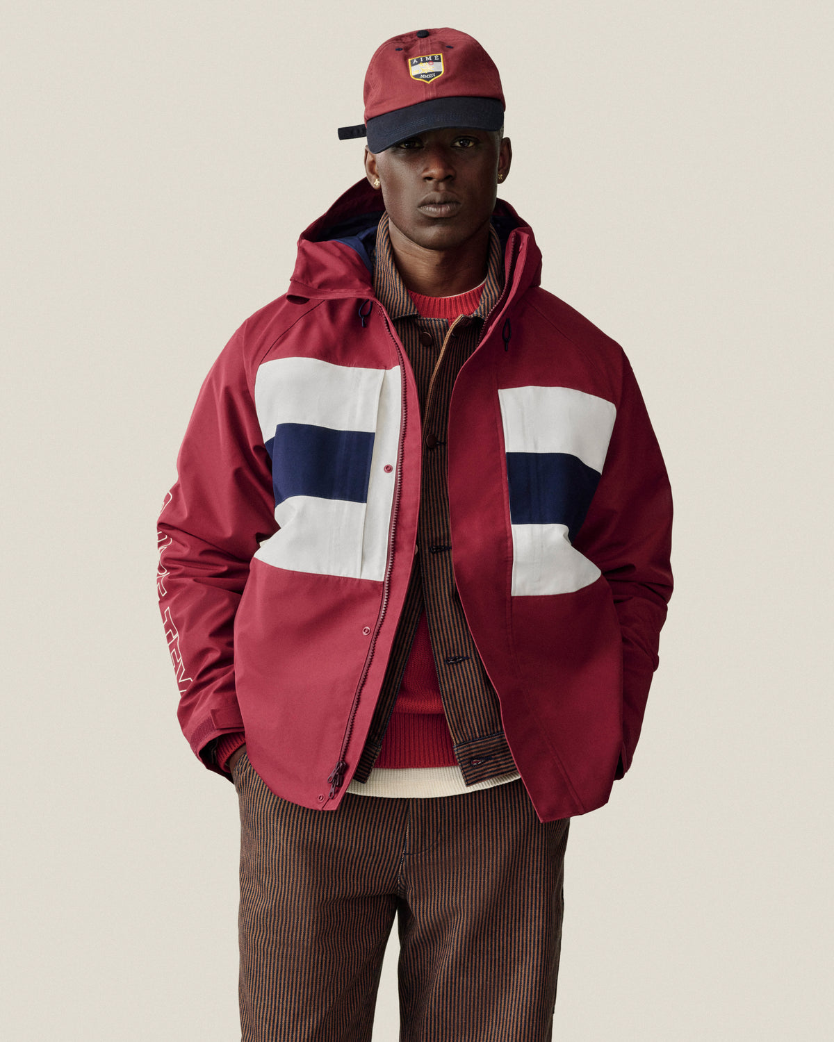 Man in maroon jacket and cap wears Hickory Stripe Painter Pant on a plain background.