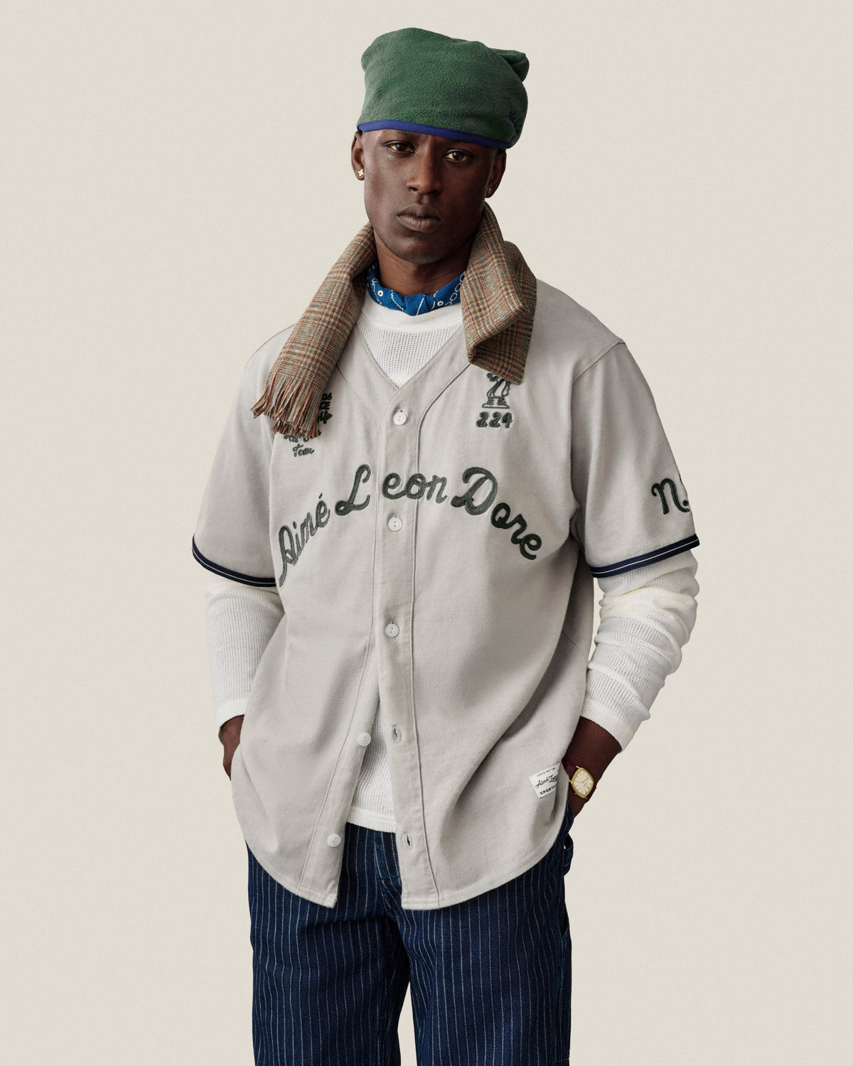 A man in a Long-Sleeve Mesh Football Tee, green beret, scarf, and pinstriped pants stands against a plain background.
