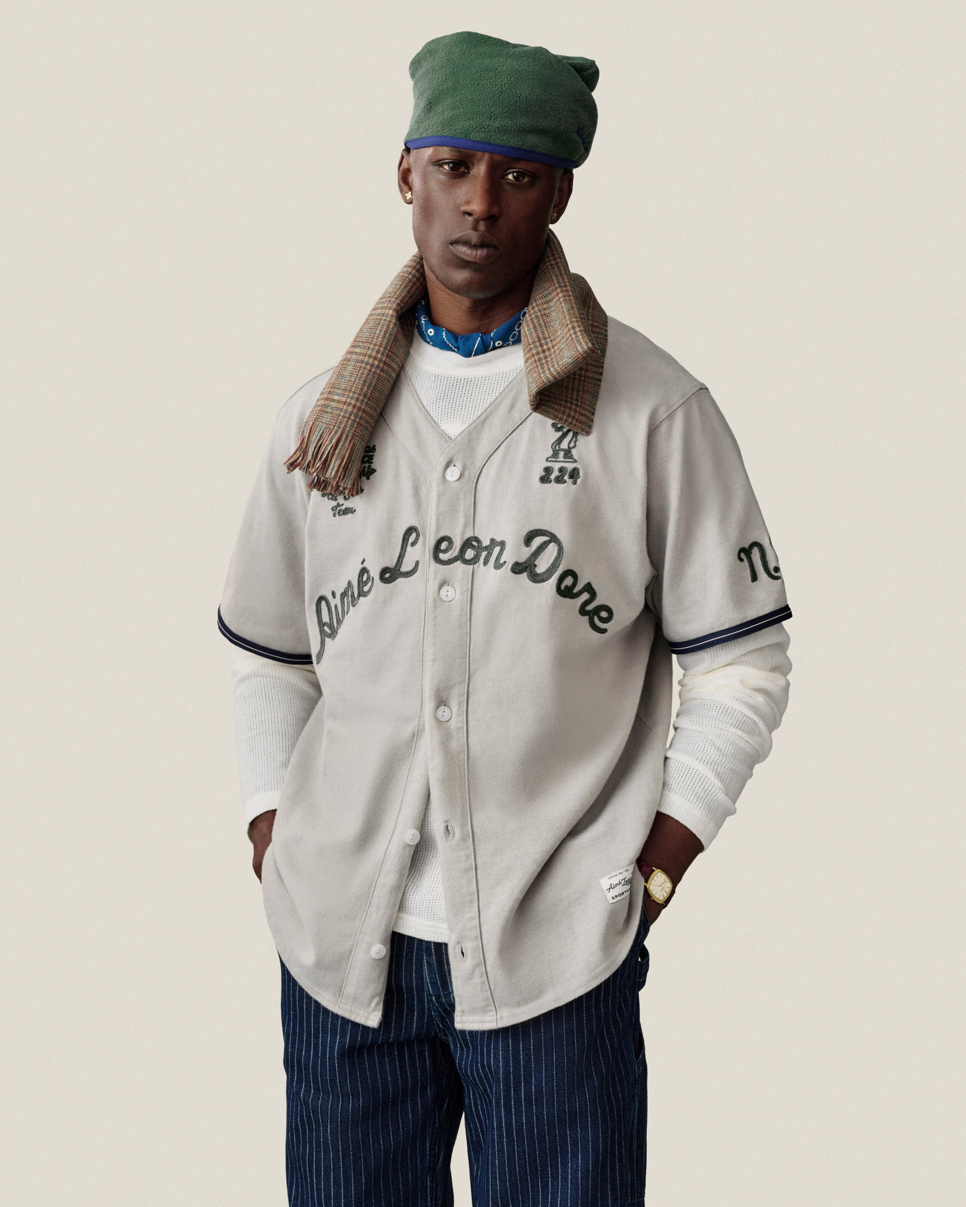 Man in Chalk Stripe Painter Pant, beige jersey, green hat, plaid scarf, and white shirt on plain background.