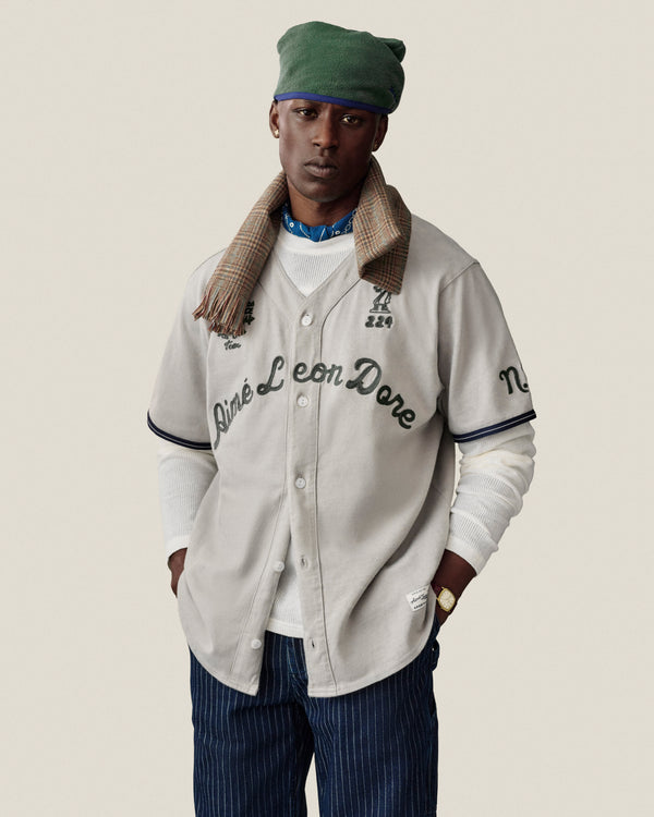 Man in Chalk Stripe Painter Pant, beige jersey, green hat, plaid scarf, and white shirt on plain background.