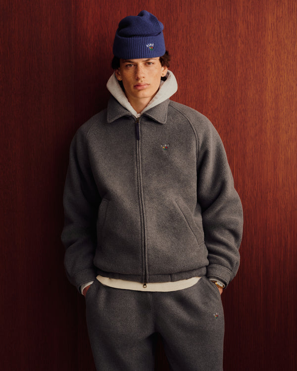Someone wearing Crest Fleece Sweatpants stands by a wood-paneled wall in a gray jacket, hoodie, and blue beanie.