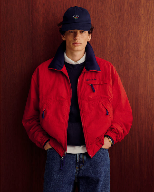 Young man in a Crest Raglan Sweater and blue jeans stands by a wooden background, hands in his pockets.