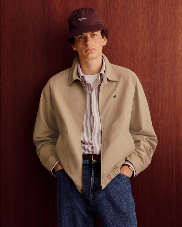 Young person in a Crest Casual Jacket, striped shirt, jeans, and maroon cap stands by a wooden wall.