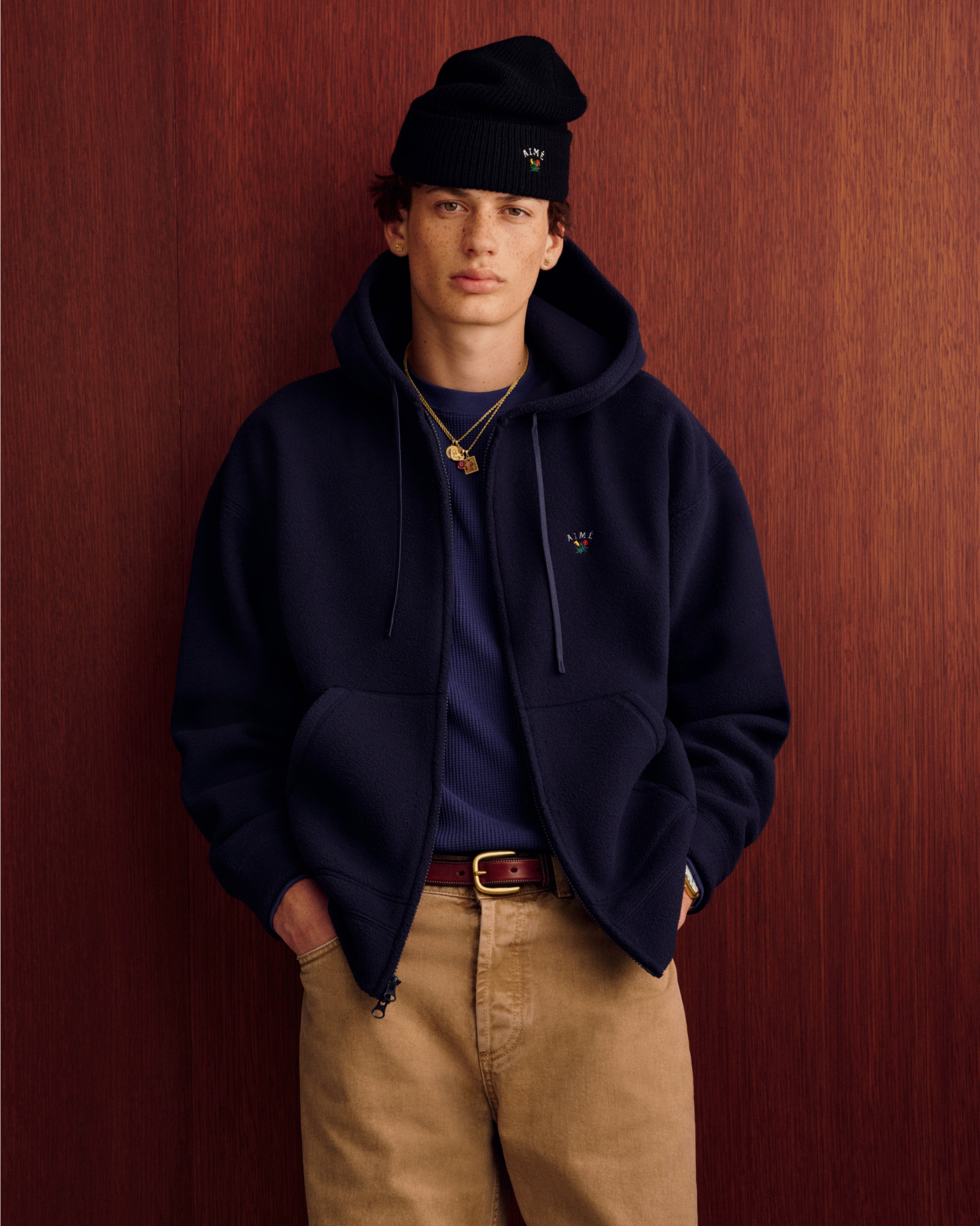 Young man in navy hoodie, Crest Merino Wool Beanie, and khaki pants stands with hands in pockets by wood panel wall.