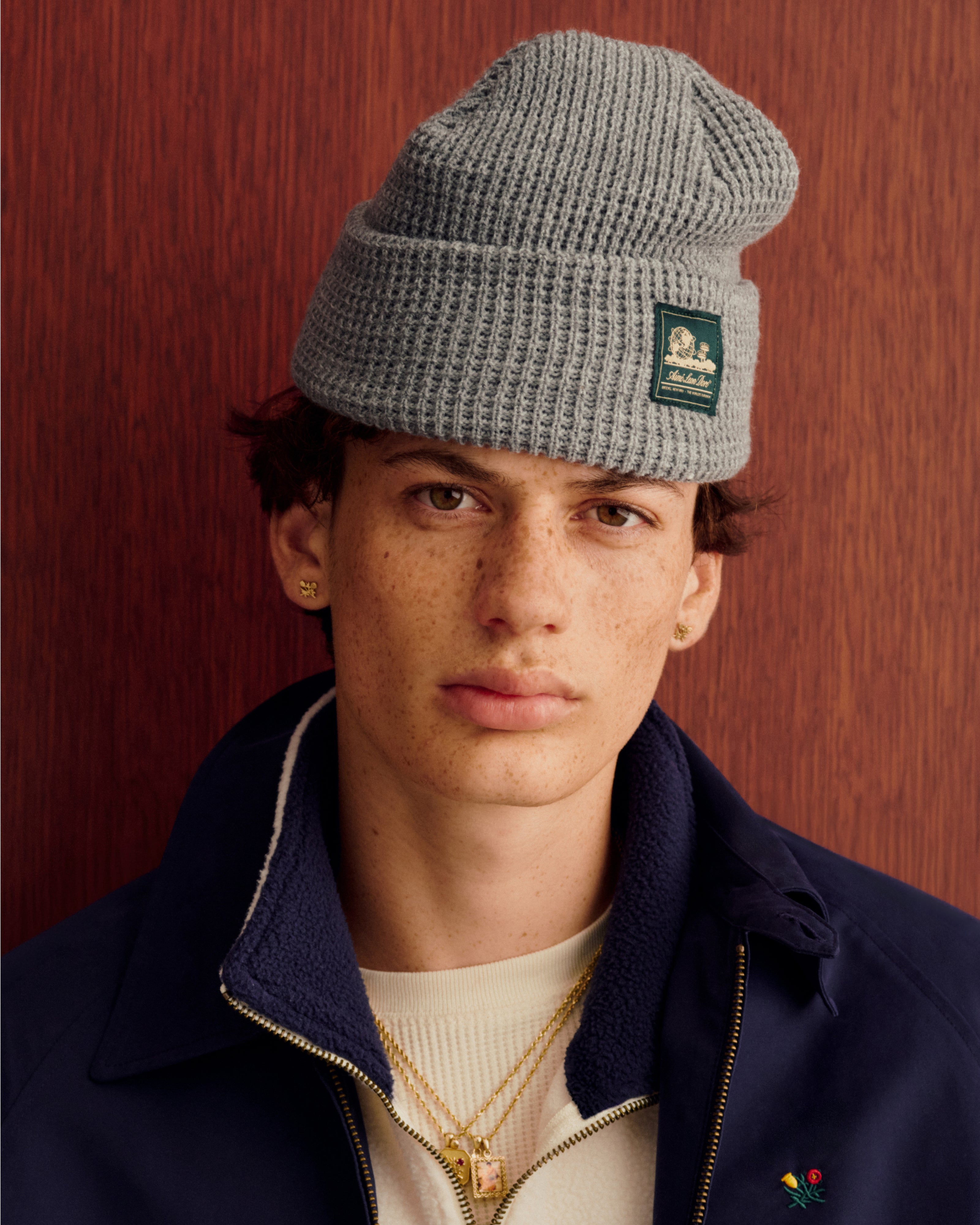 Freckled youth in gray Waffle Knit Beanie, gold jewelry, cream shirt, navy jacket, by wood panel.