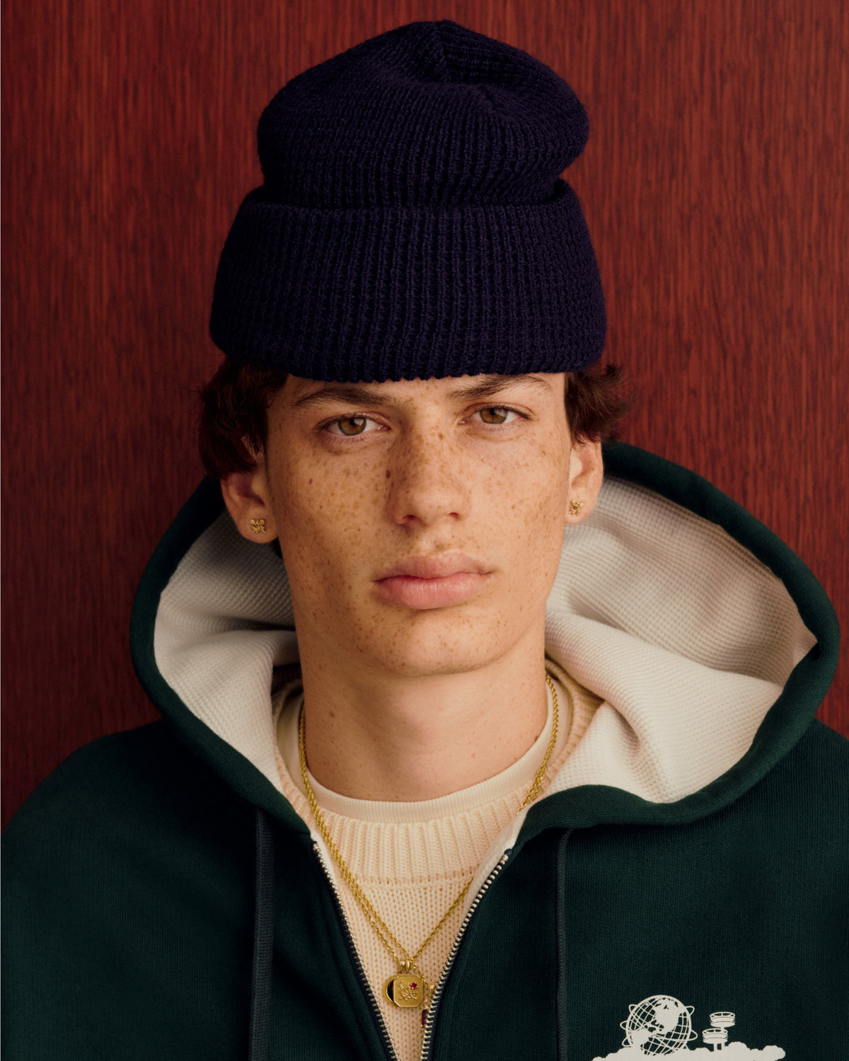 Young person in a navy Waffle Knit Beanie, green hoodie, and gold necklaces stands by a wood background.