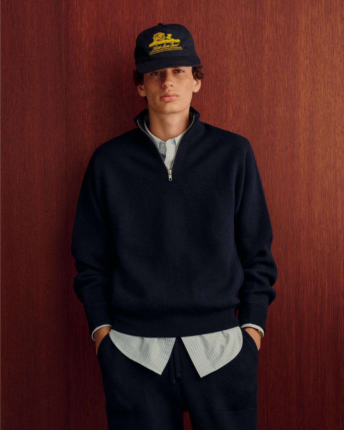 Young person wears Quarter Zip Pavilion Cashmere Pullover, striped shirt, dark cap, hands in pockets by a wooden wall.