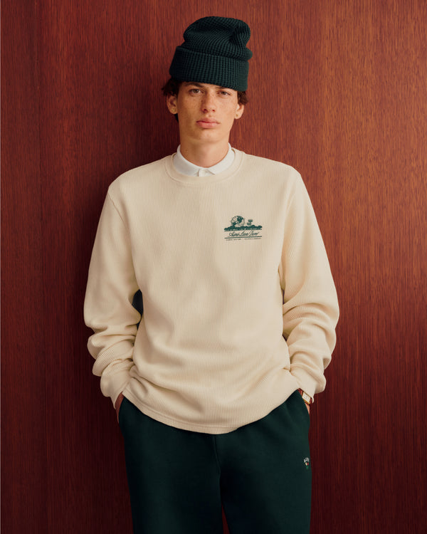 Individual in Long-Sleeve Unisphere Waffle Thermal stands with hands in pockets by a wooden backdrop.