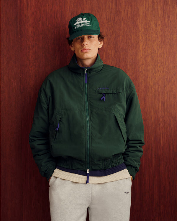 Person wearing a green jacket and Unisphere Hat, hands in pockets, gray sweatpants, against a wooden wall.