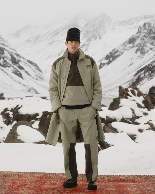 A person wearing the ALD / The North Face Jacquard Beanie stands on a red rug outdoors with snowy mountains behind.