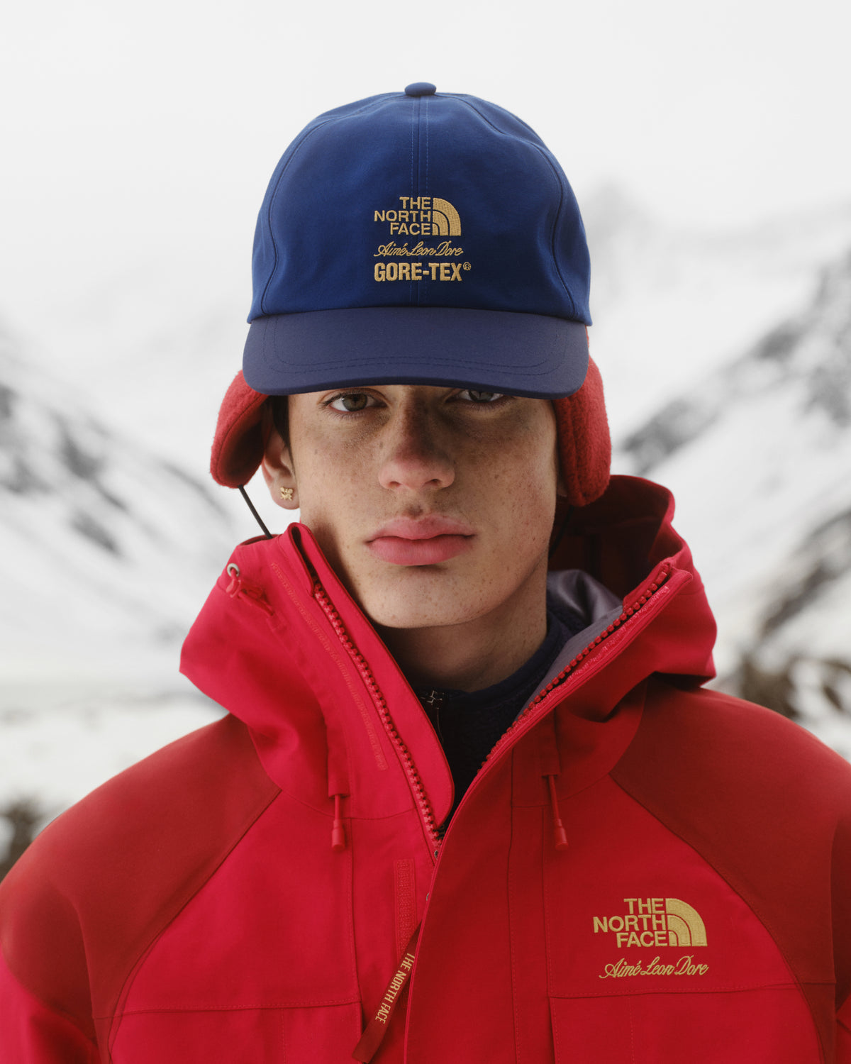 Person in red North Face jacket and ALD / The North Face GORE-TEX Hat, standing outdoors in snowy mountains.