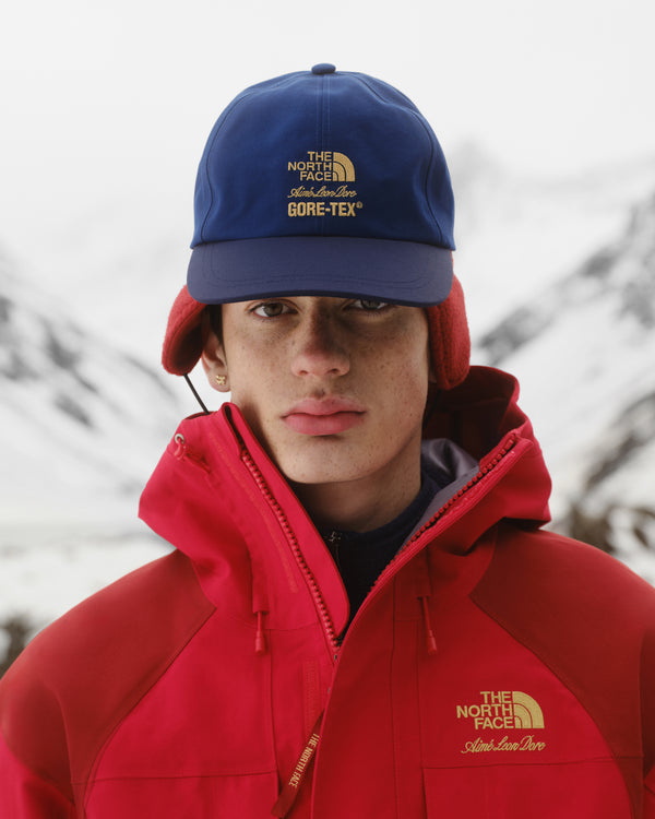 Person in red North Face jacket and ALD / The North Face GORE-TEX Hat, standing outdoors in snowy mountains.
